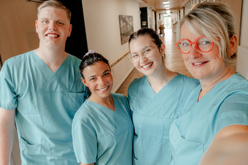 Vier Pflegekräfte in blaugrüner Funktionskleidung stehen in einem Stationsflur und blicken für ein Selfie in die Kamera im Albertinen Krankenhaus in Hamburg-Schnelsen