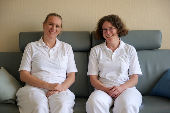 Zwei Therapeutinnen der Palliativeinheit sitzen in weißer Funktionskleidung auf einem Sofa und lächeln in die Kamera im Interdisziplinären Therapiezentrum im Albertinen Krankenhaus Hamburg Zwei Therapeutinnen der Palliativeinheit sitzen in weißer Funktionskleidung auf einem Sofa und lächeln in die Kamera im Interdisziplinären Therapiezentrum im Albertinen Krankenhaus Hamburg