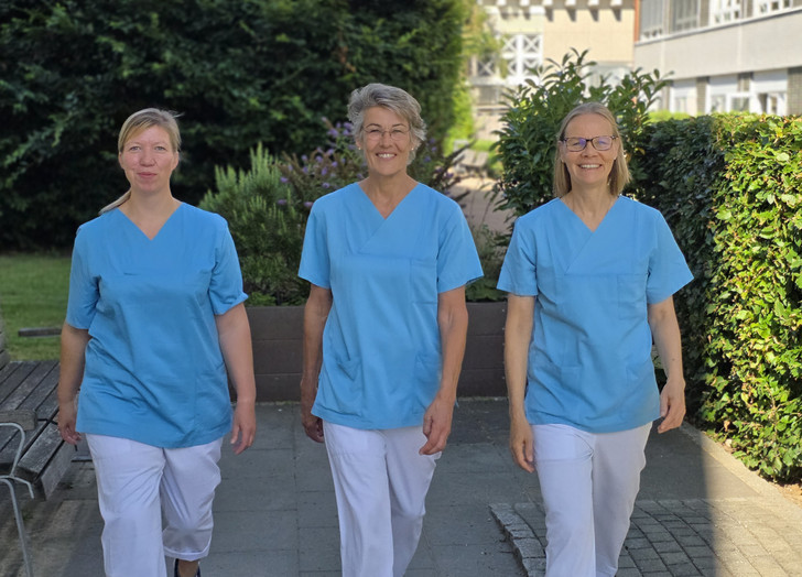 Stefanie Heldt, Leitung interdisziplinäre Therapieabteilung, läuft zwischen ihren beiden Kolleginnen aus dem Leitungsteam in blauer Funktionskleidung über das sonnige Außengelände mit Sträuchern und Bäumen des Evangelischen Amalie Sieveking Krankenhauses in Hamburg.