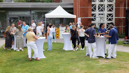 Besucherinnen und Besucher stehen um mit weißem Stoff bezogenen Stehtischen auf einer Wiese und unter einem Zeltdach beim Sommerfest 2026 auf dem Gesundheitscampus Volksdorf des Evangelischen Amalie Sieveking Krankenhauses, der Residenz am Wiesenkamp, des Feierabendhauses Volksdorf und der Albertinen Kindertagesstätte Volksdorf. 
