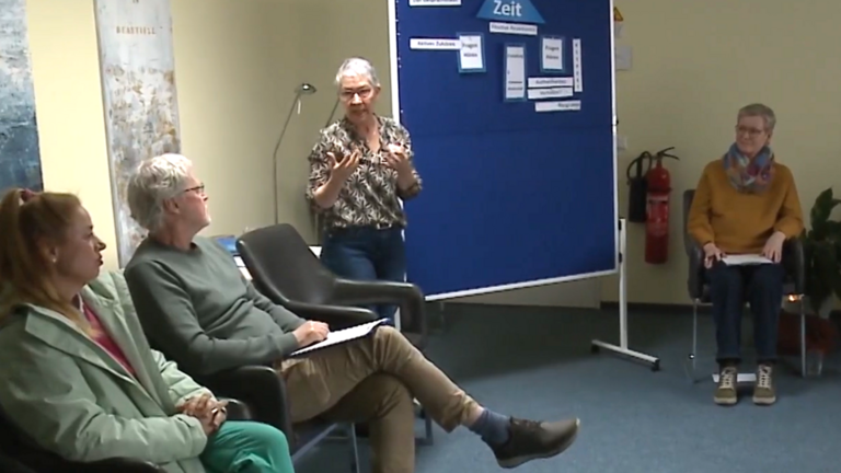 Kursleiterin Sabine Bornhöft steht an einer Pinnwand und spricht mit den im Kreis sitzenden Teilnehmenden beim Kurs für den ehrenamtlichen mobilen Hospizdienst im Albertinen Hospiz Norderstedt.