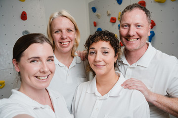 Das Team der Physiotherapie lächelt in weiß gekleidet in die Selfiekamera vor der Kletterwand im Albertinen Krankenhaus Hamburg Schnelsen