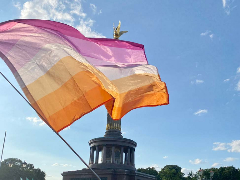 Eine LGBT-Flagge weht vor blauem Himmel und der Berliner Siegessäule zum Internationalen Tag gegen Queerfeindlichkeit