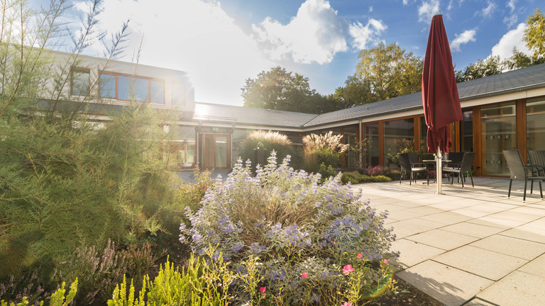 Blick auf die Vorderansicht mit Terrasse des Diakonie Hospiz Norderstedt bei blauem Himmel und Sonnenschein mit Pflanzen und Sträuchern im Vordergrund