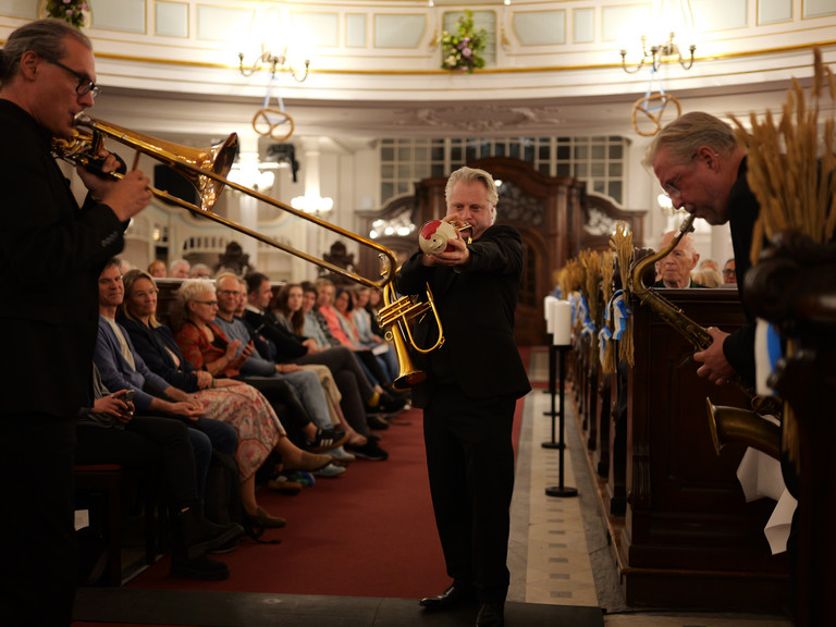 Ein Blechbläser läuft musizierend und in Begleitung von zwei anderen Bläsern des Trios "Windspiel" der NDR Bigband durch die mit Publikum besetzten Kirchengänge beim 10. Herzbrücke-Benefizkonzert im Hamburger „Michel“ der Albertinen-Stiftung.