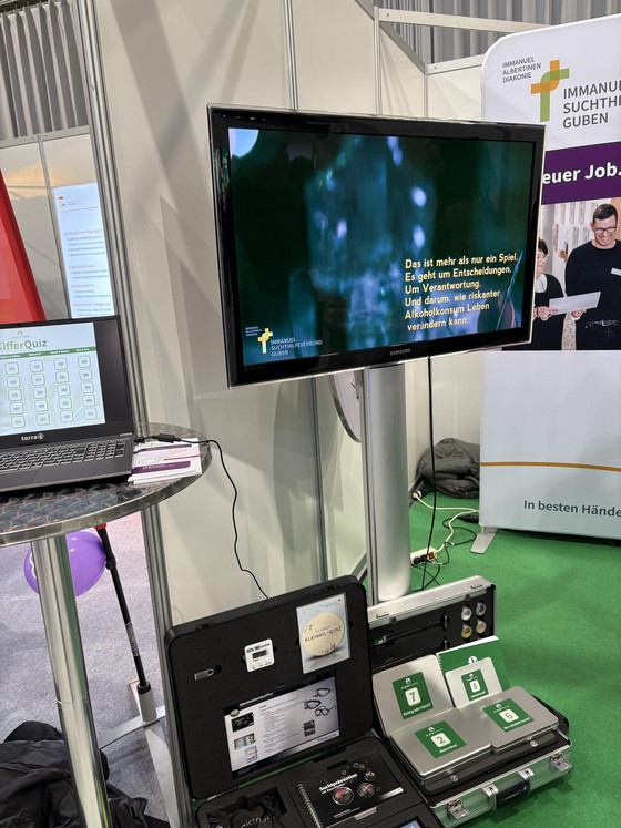 Ein großer Monitor mit Fragen zu einem Alkohol-Quiz, Koffer mit Antwortkarten und andere Materialien am Messestand des Immanuel Suchthilfeverbunds Guben auf der Ausbildungsmesse Impuls in Cottbus